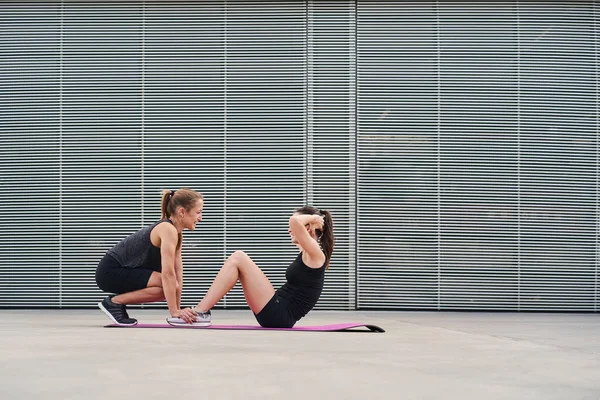 Dışarıda egzersiz yapan genç kadınlar. Bir esmer ve esmer gri metal bir duvarın önünde mekik çekiyorlar. İkisi de siyah spor giyer..