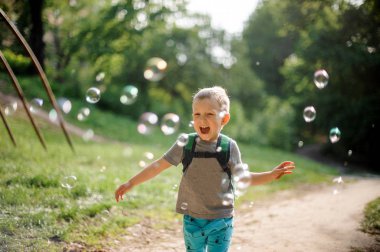 Sabun köpüğü yeşil yaz Park ile güneşli günde oynarken bir sırt çantası ile küçük çocuk gülüyor