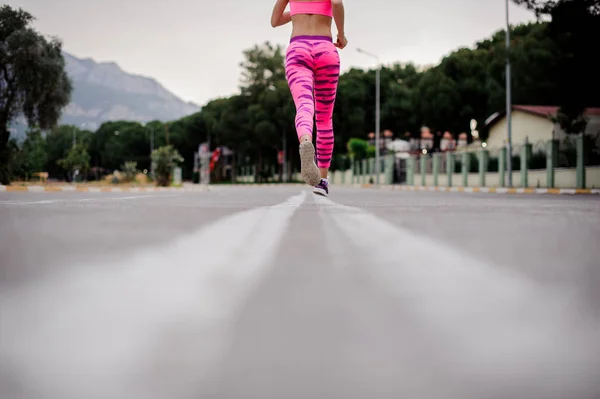Back view of slim woman legs dressed in pink sportswear running along the highway of the city