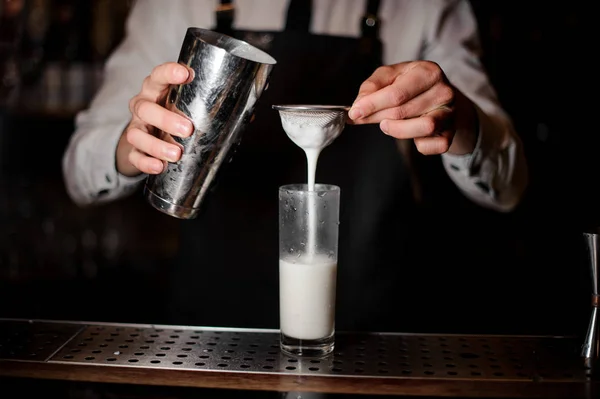 Bartender adding the alcoholic drink from a steel shaker into the drink glass on the bar counter