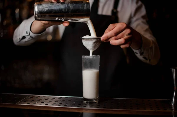 Bartender pouring an alcoholic drink from the steel shaker into a drink glass through the botler on the bar counter