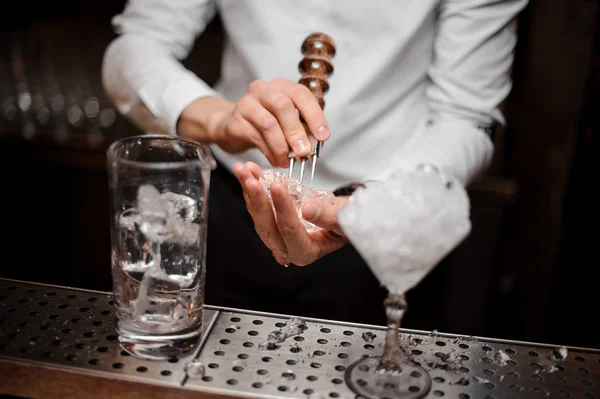 Professional bartender breaking an ice cube with the special tool to prepare a fresh cocktail at the steel bar counter