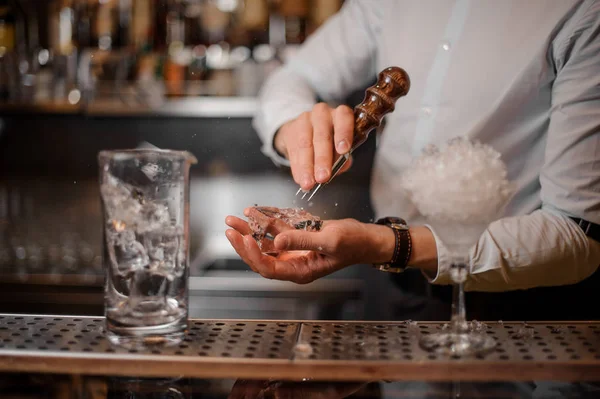 Professional bartender breaking an ice cube with the special tool to prepare a delicious cocktail at the steel bar counter