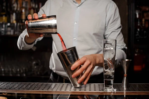 Bartender mixing in the steel cocktail shaker a transparent red alcoholic drink on the bar counter on the blurred background