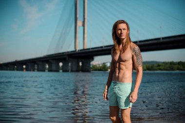 İnce bir genç adam arka plan mavi nehir ve köprü üzerinde yaz gününde ayakta portresi