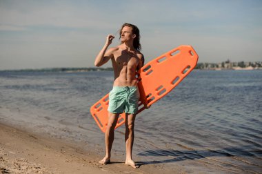 Mavi gökyüzü turuncu hayat kurtaran ekipman ile kum üzerinde duran yakışıklı seksi cankurtaran