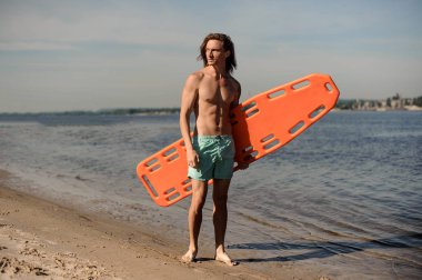 Mavi gökyüzü turuncu hayat kurtaran ekipman ile kum üzerinde duran genç seksi plaj cankurtaran