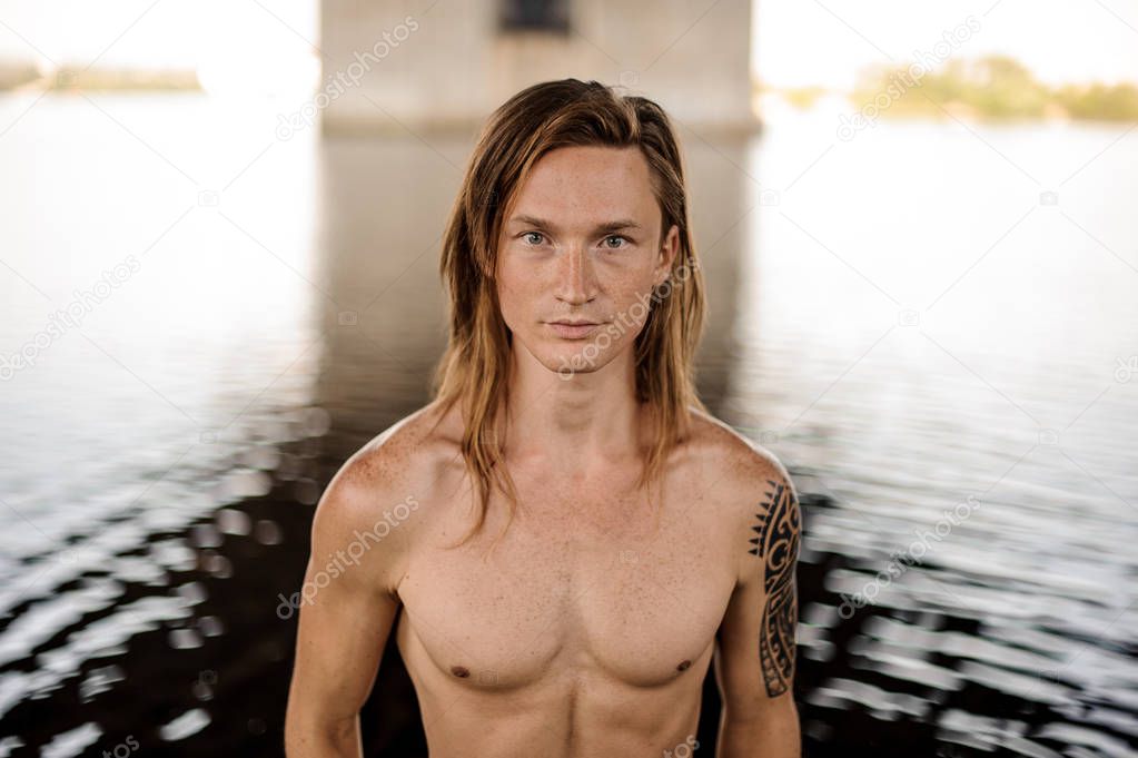 Portrait of young handsome long and red-haired man with tattoo on the shoulder standing in water