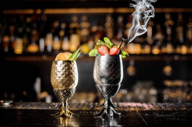 Two cocktails in the steel pineapple form cup with strawberries, mint and smoking cinnamon and with berries and orange in the bar clipart