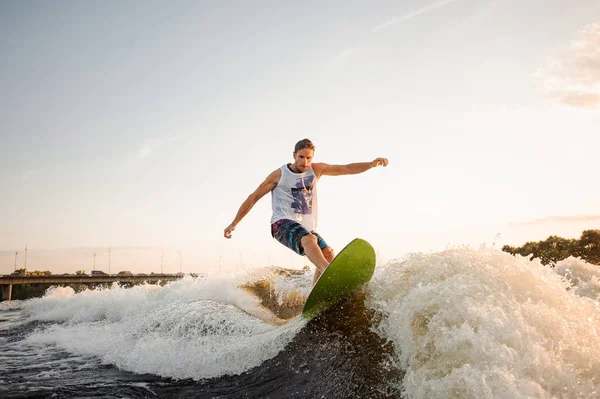 Active and young man riding on green wakesurf down the river waves against clear sky on hot summer day