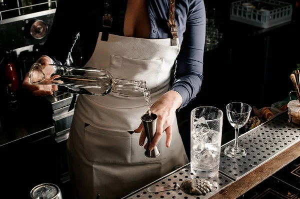 Female bartender in the white apron pouring an alcohol from the bottle into the measuring cup at the bar counter