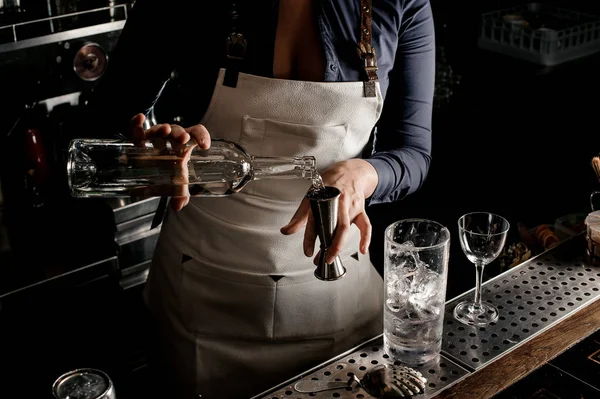 Female bartender in the white apron pouring an alcoholic drink from the bottle into the measuring cup at the bar counter