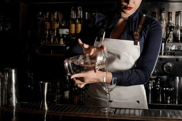 Female bartender in the white apron pouring an alcoholic drink from the measuring cup into the cocktail glass at the bar counter