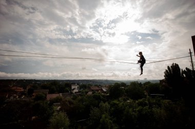 Tek ayak üstünde slackline ip ağaçlar ve berrak gökyüzü arasında evlerin zemin üzerine oturan genç aktif sarışın kadın