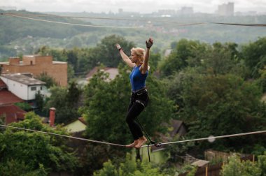 Ağaçlar ve berrak gökyüzü arasında evlerin zemin üzerine bükme diz slackline ip üzerinde duran genç kadın
