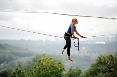 Arka plan ağaç ve berrak gökyüzü arasında evlerin slackline ipe üzerinde oturan yan görünümü genç sarışın kadın
