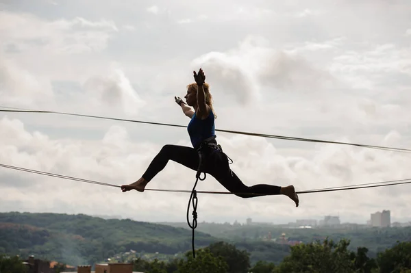 Young blondie woman trying to sit on twine on the slackline rope on the background of houses among trees and clear sky