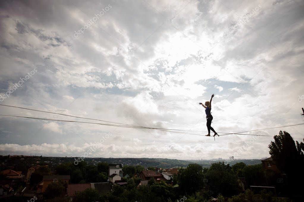 Young blondie woman making steps on the slackline rope on the background of houses among trees and clear sky