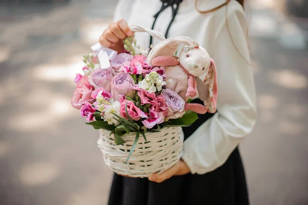 Colegiala sosteniendo una linda canasta de mimbre llena de flores de color rosa brillante decoradas con un juguete — Foto de Stock