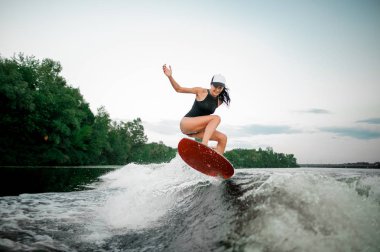 Genç gülümseyen esmer kadın Motorbot ağaçlar arka plan üzerinde yüksek dalga üzerinde turuncu wakesurf atlama
