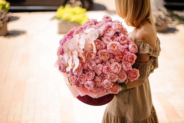 Young girl in beige dress holding in her hands a huge bouquet of peony roses decorated with pink orchids