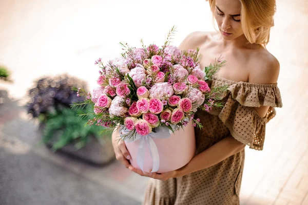 年轻漂亮的女孩穿着米色的礼服 手里拿着一盒嫩粉色的玫瑰和牡丹 点缀着绿色的树枝 — 图库照片
