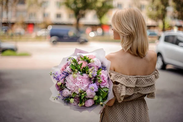 緑の葉で飾られた柔らかいピンクと紫の花のブーケを手に持ったベージュのドレスで背面ビューのブロンドの女の子 — ストック写真