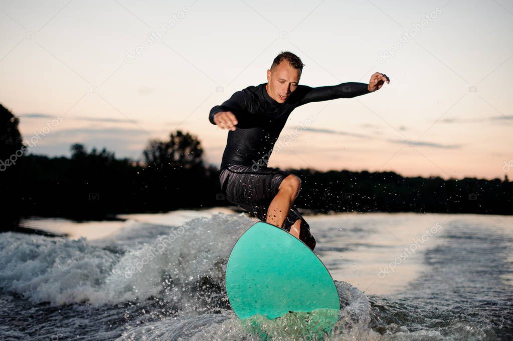 Active wakesurfer in black swimsuit jumping on a wake board riding down the river at the evening