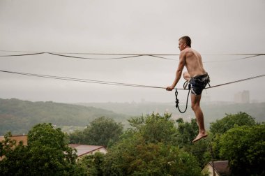 Cesur adam yağmurlu yaz gününde bir slackline gökyüzü ve ağaçlar karşı denge tutmak