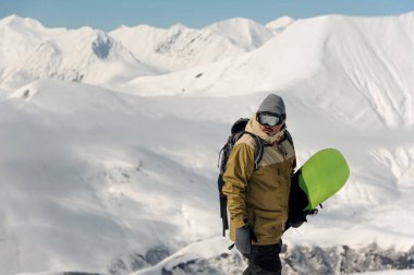 adam kayak ekipmanları ve gözlük yeşil bir snowboard üzerinde arka plan karşı karlı dağlar tutuyor