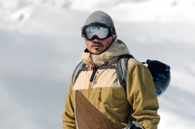 karlı bir arka plan üzerinde güneşin kayak ekipmanları ve gözlük adamla ahşap snowboard onun elinde duruyor