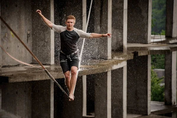 Strong and brave man balancing on a slackline against the background of high empty building on summer day