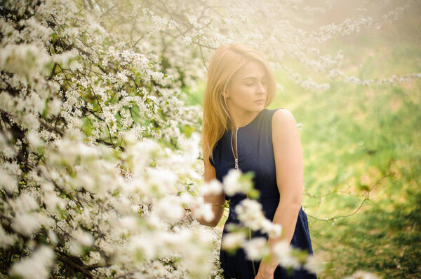Attractive blonde woman standing on the blurred background of a white blooming cherry tree on sunny day