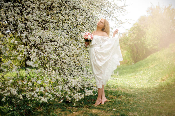 Romantic full height portrait od a blonde woman in white dress with a bouquet standing on tiptoe near the blooming cherry tree