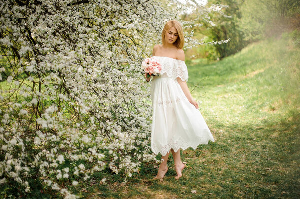 Romantic full height portrait od a blonde woman in white dress with a bouquet standing near the blooming cherry tree