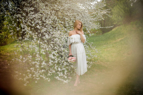 Tender full height portrait od a blonde woman in white dress with a bouquet standing near the blooming cherry tree on misty day
