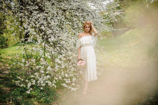 Full height portrait od a blonde woman in white dress with a bouquet standing near the blooming cherry tree on misty day