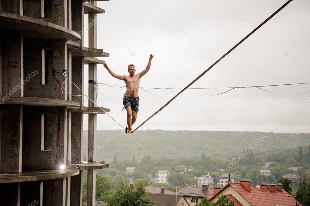 Brave man balancing on a slackline high against empty building and sky on rainy summer day