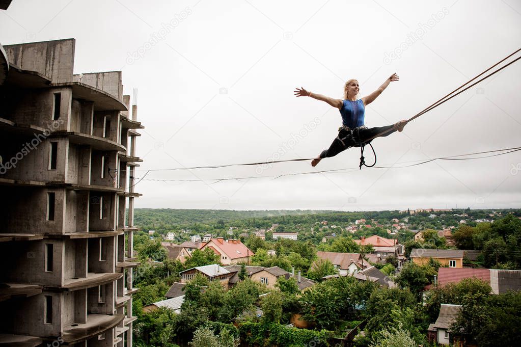Brave and happy woman balancing on a slackline against the background of high empty building on summer day