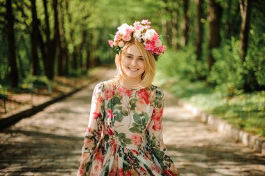 Gülümseyen Park'ta bir çiçek elbise ve yeşil ağaçlar arka plan üzerinde kafa onu ayakta üzerinde pembe çelenk giyinmiş sarışın kadın