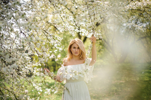 Romantic portrait of a happy blonde woman in white dress near the blooming cherry tree