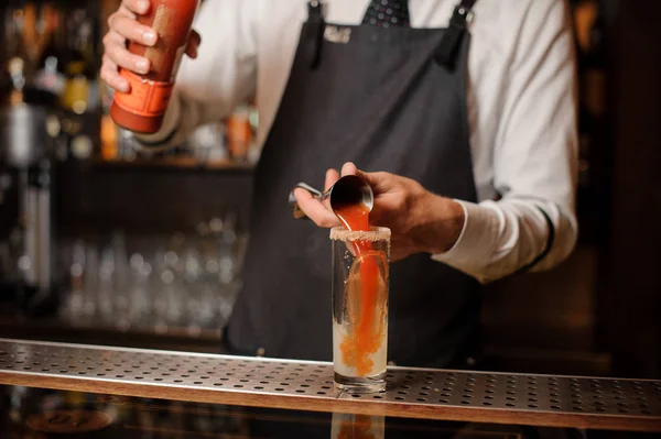 Bartender in the white shirt and dark apron pourring a bloody mary into the cocktail glass with an ice cube on the bar counter