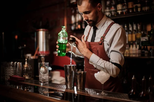 酒保在白色衬衣和棕色皮革围裙 Pourring 一个喝酒也是从测量杯子入钢振动筛和拿着一个绿色瓶 — 图库照片
