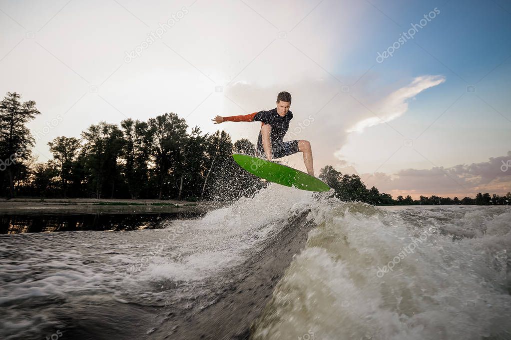 Young attractive man jumping on the wakeboard on high wave of motorboat on the background lakeside