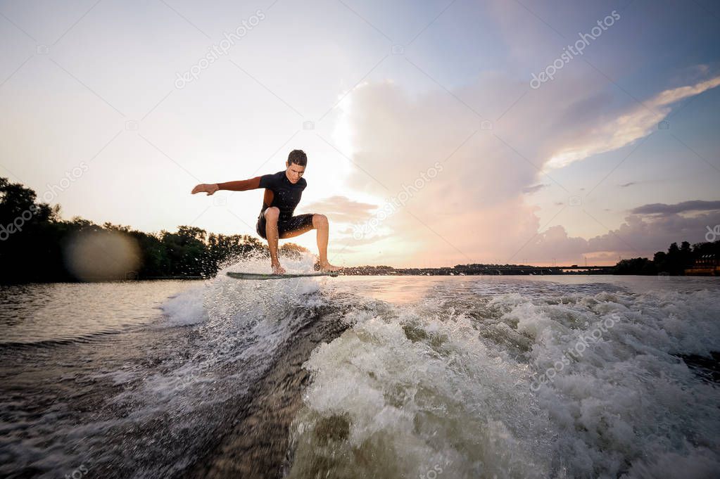 Young attractive man riding on the wakeboard on high wave of motorboat on the background of a lakeside