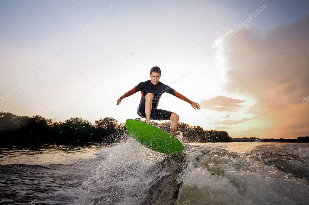 Young attractive man making jump on the green wakeboard on high wave of motorboat on the background of a lakeside