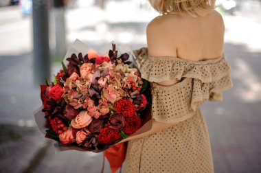 Kırmızı tonlarda romantik bir buket çiçek tutan bej içinde sarışın genç kadın arkadan görünüm