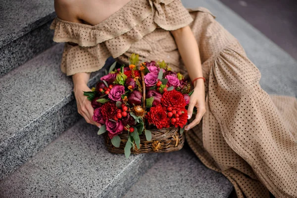 Girl in beige floor-length dress sitting on concrete stairs with a wicker basket of flowers