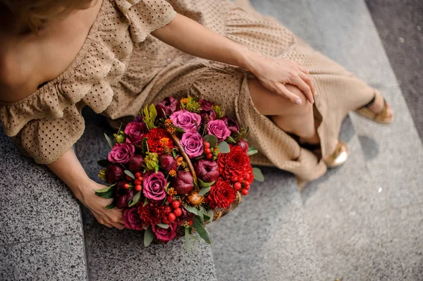Elegant woman in beige floor-length dress sitting on concrete stairs with a wicker basket of flowers
