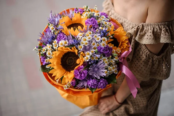 穿着米色连衣裙的女孩捧着一束鲜艳的紫色花朵 向日葵和 Chamomiles 饰着蝴蝶结 — 图库照片